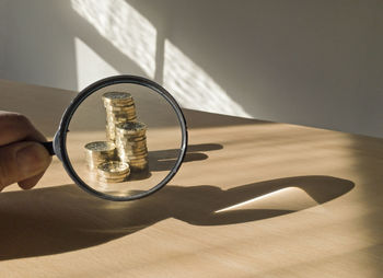 Cropped hand of person holding magnifying glass against coins on table