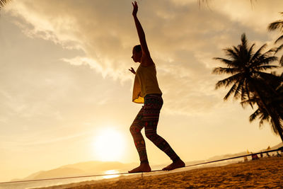 Woman slacklining on rope at beach against sky during sunset