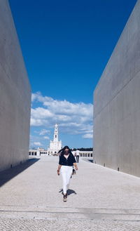 Full length of woman walking on footpath amidst wall against blue sky