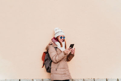 Woman wearing warm clothing using smart phone while standing against wall