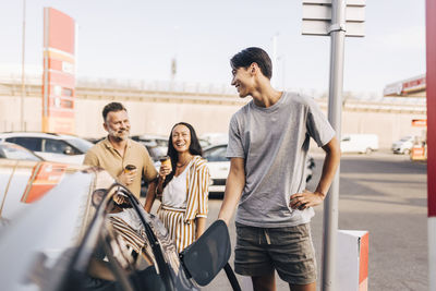 Happy young man charging car with electric plug while talking with parents at station