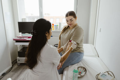 Doctor tying band on patient's arm sitting on bed in clinic