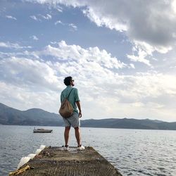 Rear view of man standing on pier over lake against cloudy sky