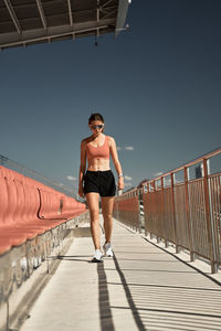 Full body of determined young sportswoman in activewear and sunglasses walking along tribune of stadium against blue sky