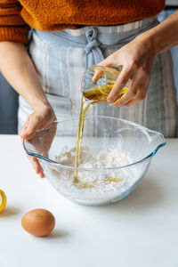 Crop female pouring olive oil from glass into large bowl with flour while preparing dough at white table in kitchen