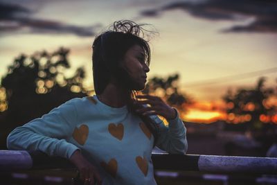 Young woman leaning on railing against sky during sunset