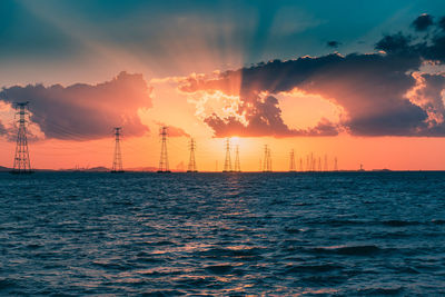 Electricity pylons by sea against sky during sunset