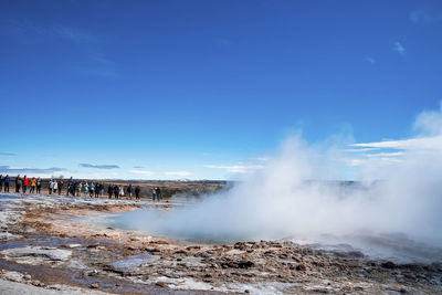 Tourists exploring strokkur geyser eruption in valley against blue sky