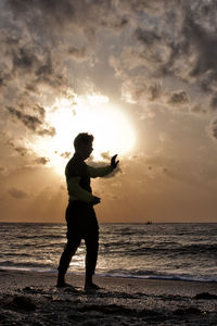 Silhouette man practicing martial arts at beach against sky during sunset