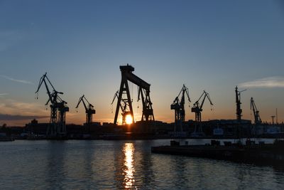 Silhouette cranes at harbor against sky during sunset