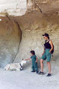 Family of tourists a mother with a backpack in hat and a son stand on mountain near rock next goat