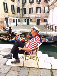 Side view of gondolier sitting on chair by canal in city