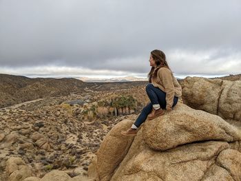 Side view of woman sitting on rock against sky