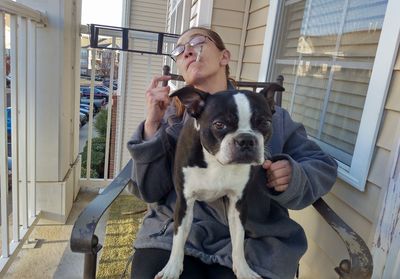 Woman smoking while sitting with boston terrier on chair at porch