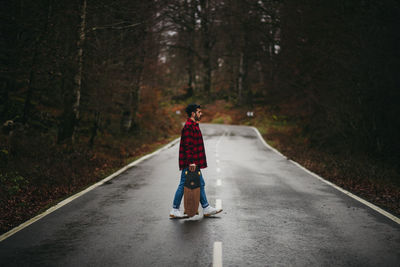 Side view of young stylish man in casual wear walking on asphalt road with skateboard in hand on autumn day