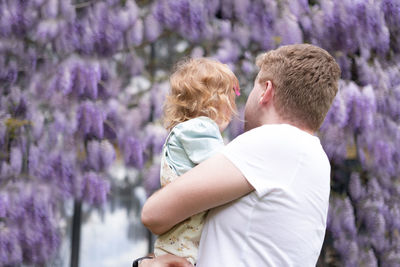 Father holding baby girl,daughter outdoor looking at glucinum flowers,purple,very peri wistaria tree
