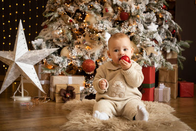 Cute baby girl holding bauble sitting against christmas tree at home
