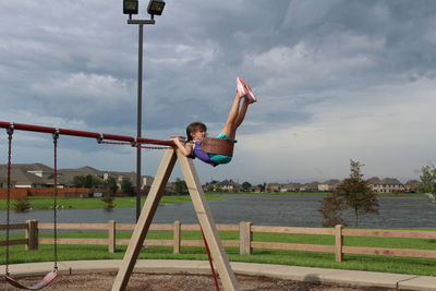 Girl enjoying swing at playground by lake against cloudy sky