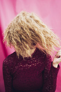 Unrecognizable female model in red dress touching blond wavy hair on pink background in studio