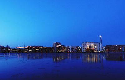 Illuminated buildings by river against blue sky at dusk