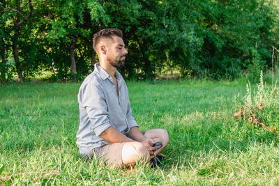 Portrait of young handsome european man in casual clothing sitting on a grass in summer park.