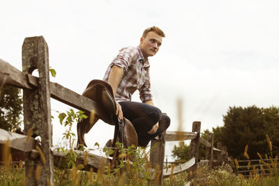 Low angle view of rancher sitting on wooden fence against sky