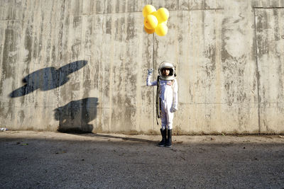 Girl wearing space suit holding yellow balloon standing against wall during sunny day
