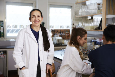 Portrait of confident mature female chemistry teacher standing by students in laboratory at university