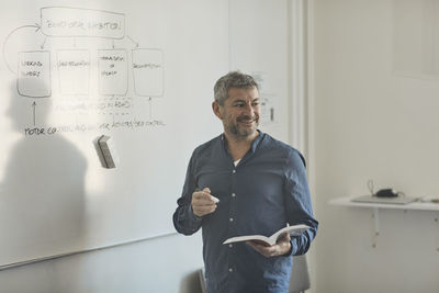 Smiling male teacher with book standing by whiteboard in classroom