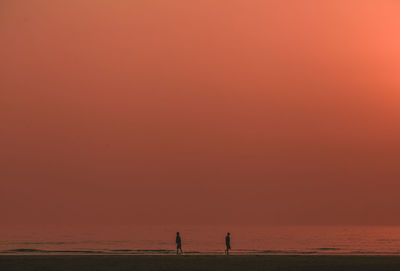 Silhouette people on shore at beach against orange sky during sunset