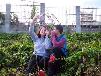 Young woman taking selfie with friend while sitting amidst plants against fence