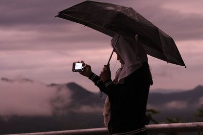 Woman photographing while holding umbrella against cloudy sky during rain