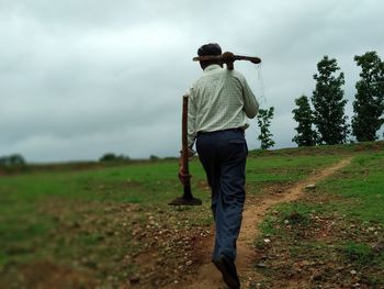 Rear view of man carrying pick axe and shovel on field