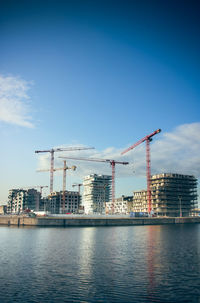 Cranes by river and buildings against sky