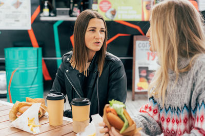Two young modern european girlfriends are chatting and eating burgers in a street market.