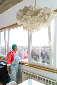 Woman with short dyed green hair looking out window overlooking city with buildings while standing in light room at home