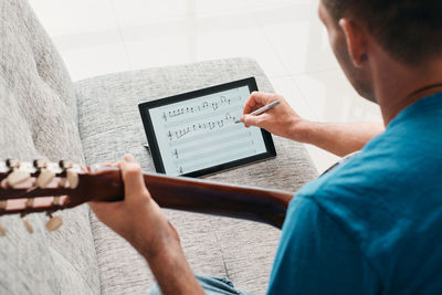Rear view of man holding guitar writing on digital tablet on sofa at home