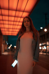 Portrait of woman holding mobile phone while standing on footpath under illuminated lights