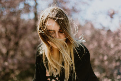 Beautiful young woman looking down with tousled hair standing against trees