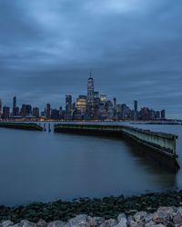 Modern buildings by river against cloudy sky at dusk