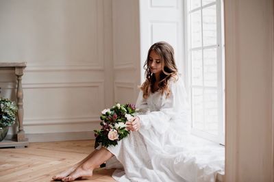 Bride holding bouquet while sitting at window sill in room