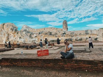 People sitting on rock against cloudy sky