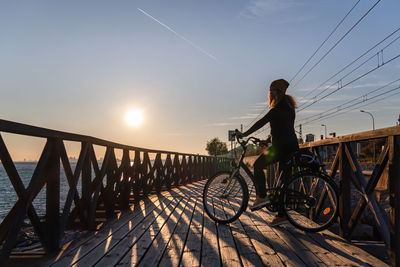 Woman riding bicycle on bridge against sky during sunset