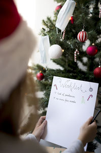Cropped image of girl writing on paper by christmas tree at home