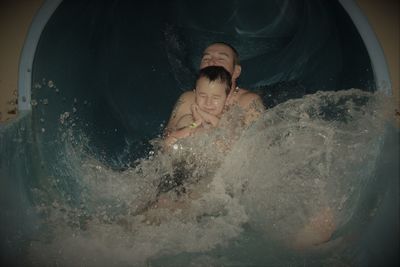 Shirtless man with son sliding in water slide