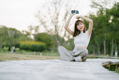 Young woman listening music while sitting on land against trees