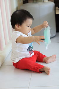 Cute baby girl holding surgical mask sitting on floor at home