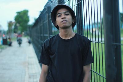 Portrait of young man wearing hat standing by fence