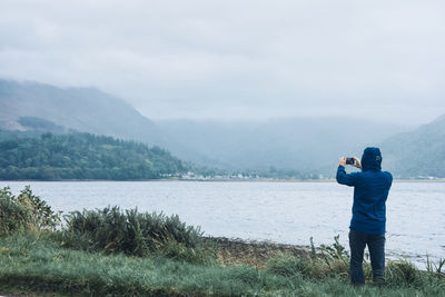 Rear view of a man standing by a lake taking a photo with a mobile phone of a dramatic landscape.