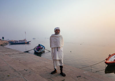 Varanasi, india - february, 2018: elderly indian man wearing traditional white clothes standing on river bank with fishing boats on water in early foggy morning